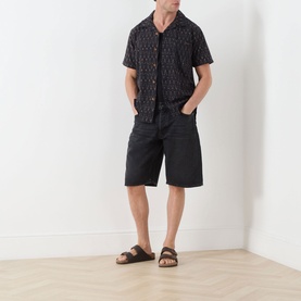 Model wearing Nudie Jeans Night Dune shorts, styled with a dark patterned short-sleeve shirt and brown leather sandals.