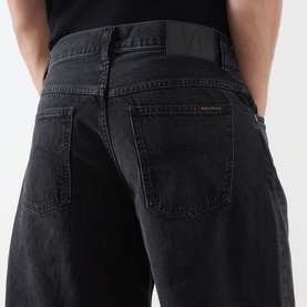 Model wearing Nudie Jeans shorts in Night Dune black denim, rear view showing back pockets and orange logo tab detail.