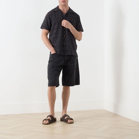 Model wearing Nudie Jeans Night Dune shorts, styled with a black printed short-sleeve shirt and brown leather sandals.