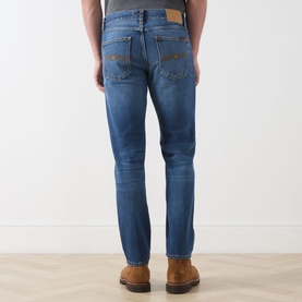 Model wearing Nudie Jeans Gritty Jackson Straight-Leg Jeans in Sun Set, showing rear view with tobacco stitching, logo badge and faded mid-blue wash.