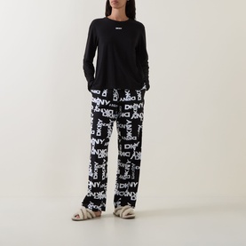 Model wearing DKNY Black Logo Plaid Pyjamas; black long-sleeve top and logo-print trousers, styled with cream fluffy slippers.