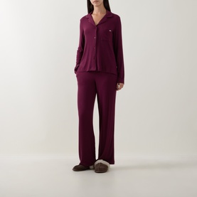 Model wearing DKNY Maroon Banner pyjamas in soft viscose blend, long-sleeve button-up top and wide-leg trousers, styled with dark slippers