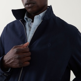Model wearing a navy zip-up harrington-style jacket with embroidered logo, styled over a light blue striped shirt