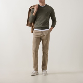 Model wearing Hugo Boss Orange light pastel brown cotton chino trousers, styled with olive crew-neck jumper, white tee and white trainers.