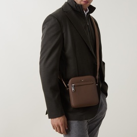 Model carrying Hugo Boss RayNS Logo Reporter Bag in medium brown with silver-tone hardware, worn crossbody over dark blazer and grey trousers.