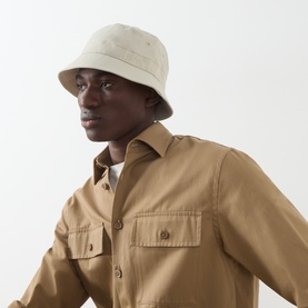 Model with Hugo Boss Finnian Logo Bucket Hat in light beige, featuring narrow stitched brim, styled with a tan cotton overshirt
