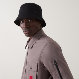 Model with Hugo Boss Finnian Logo Bucket Hat in black, featuring narrow stitched brim and rubberised logo, styled with a taupe overshirt.