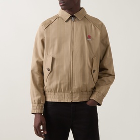 Model wearing Hugo medium beige casual jacket with embroidered chest logo, elasticated hem, and snap-button pockets, styled with black trousers.