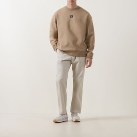 Model wearing Hugo Beige Overflow cotton crew neck sweatshirt with small logo patch, styled with light grey trousers and white trainers.