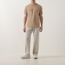 Model wearing Hugo medium beige cotton crew neck T-shirt with tonal logo detail, styled with light grey trousers and white trainers.