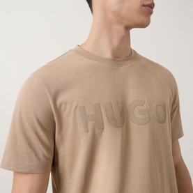Model wearing Hugo medium beige cotton crew neck T-shirt with tone-on-tone embossed logo lettering across the chest.