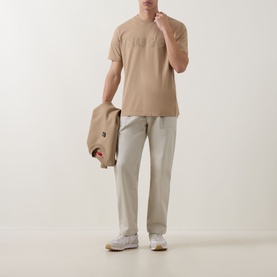 Model wearing Hugo medium beige cotton crew neck T-shirt, styled with light grey trousers and white trainers, carrying a beige jacket.