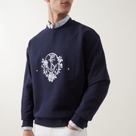 Model wearing Hugo dark blue cotton crew neck sweatshirt with white anchor and floral embroidery, styled with a light blue shirt underneath.
