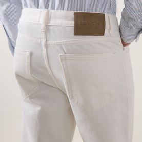 Model wearing Hugo 3049 Jeans in natural white, close-up of back pockets with tan leather logo patch, styled with blue striped shirt