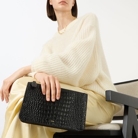 Model carrying Toteme Threefold Croco-Embossed Clutch in black croco-embossed leather with gold-tone hardware, styled with cream knit and yellow