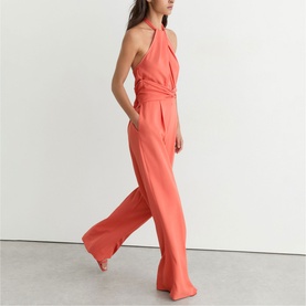 Model wearing Reiss coral pink halterneck jumpsuit with twist-front bodice and wide-leg trousers, styled with block-heel sandals.