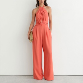 Model wearing Reiss coral pink halterneck wide-leg jumpsuit with crossover twist front detail, styled with a woven clutch bag