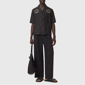 Model wearing All Saints jet black short-sleeve shirt with embroidered chest patches, styled with matching wide-leg trousers and brown sandals.
