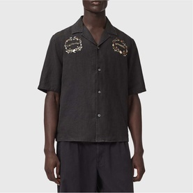 Model wearing All Saints jet black short-sleeve shirt with gold embroidered chest detail, styled with matching black shorts.