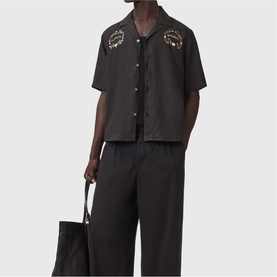 Model wearing All Saints jet black short-sleeve shirt with gold embroidery, styled with black wide-leg trousers, carrying a black tote bag.