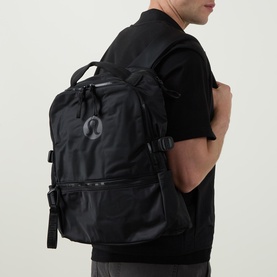 Model carrying Lululemon New Crew Backpack 22L in Black/Graphite Grey, worn on back, with top carry handle and front buckle straps visible.
