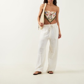 Model wearing Farm Rio Strelitzia Printed Handkerchief Bikini Top in Light Blush with white linen trousers and flat sandals, carrying a tote bag.