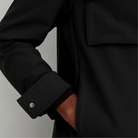 Model wearing Reiss black coat, close-up of cuff detail with silver button and chest pocket visible, hand in pocket
