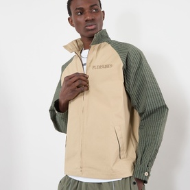 Model wearing Pleasures beige casual jacket with green corduroy contrast sleeves and stand collar, styled with white tee and green trousers