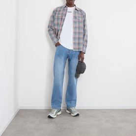 Model wearing Pleasures pink plaid cotton casual shirt with white tee, light blue jeans and grey trainers, holding a dark cap.