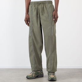 Model wearing Pleasures Sound Logo Track Pants in olive, tapered-leg nylon trousers with side piping and elasticated waist, styled with chunky