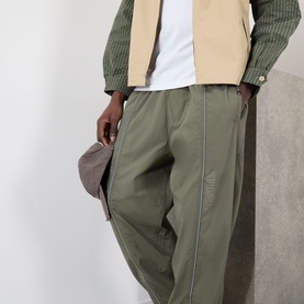 Model wearing Pleasures Sound Logo Track Pants in olive, tapered leg with contrast piping. Styled with a striped shirt and camel gilet. Holding a