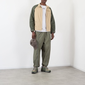 Model wearing Pleasures Sound Logo Track Pants in olive, tapered nylon fit with piping detail, styled with a two-tone jacket and trail trainers.