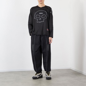 Model wearing Pleasures black nylon casual trousers with contrast stitching, styled with a black skull-print long-sleeve top and chunky trainers.