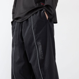 Model wearing Pleasures black nylon casual trousers with contrast piping, embroidered logo, and elasticated waist, styled with a black oversized tee.