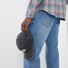 Model wearing Pleasures indigo jeans in relaxed fit with embroidered back pocket detail, styled with plaid shirt, holding dark cap.