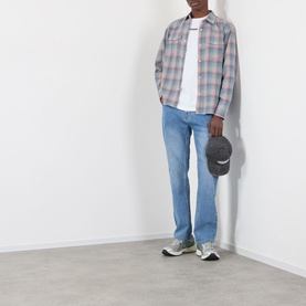 Model wearing Pleasures indigo jeans with a grey check overshirt, white tee, and grey trainers, holding a cap.