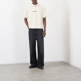 Model wearing Pleasures Cable Logo Polo Shirt in ecru with dropped shoulders and chest logo, styled with dark wide-leg jeans and brown shoes.