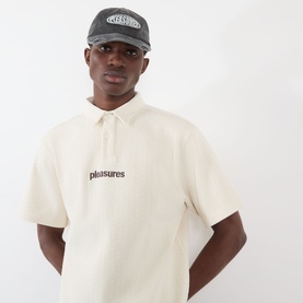 Model wearing Pleasures Cable Logo Polo Shirt in ecru with cable-knit texture and chest logo, styled with a grey cap.