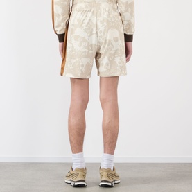 Model wearing Pleasures ecru shorts with abstract print, styled with matching top, white socks and gold trainers, seen from behind.