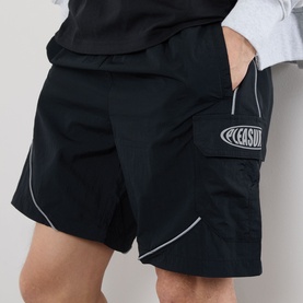 Model wearing Pleasures black nylon shorts with contrast piping, cargo-style side pocket and reflective logo patch.