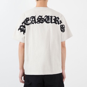 Model wearing Pleasures oatmeal cotton T-shirt, back view showing large black Gothic-style arch logo print across shoulders.