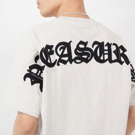 Model wearing Pleasures oatmeal cotton T-shirt, showing back detail with large black Gothic-style brand lettering across shoulders.