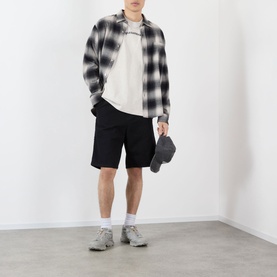 Model wearing Pleasures oatmeal cotton T-shirt, styled with black check overshirt, black shorts, and grey trainers, carrying a dark pouch bag.