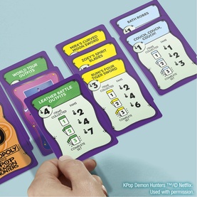 K-Pop Demon Hunters Monopoly Deal card game, multi-coloured cards fanned out on blue surface, hand placing a card