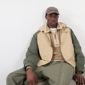 Model with Pleasures Bubble Rip Logo Baseball Cap in brown, curved visor, embroidered bubble logo, styled with beige gilet and olive jacket.