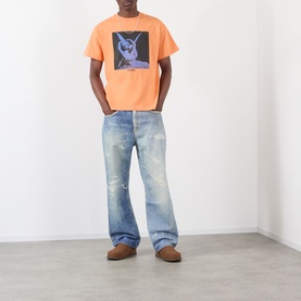 Model wearing Pleasures orange graphic T-shirt with blue distressed jeans and tan suede shoes