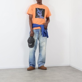 Model wearing Pleasures orange graphic print T-shirt with blue jeans tied at waist, dark cap, and brown shoes