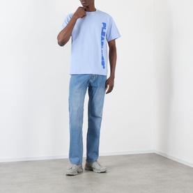 Model wearing Pleasures Buzzed Tee in sky blue with vertical tonal logo, styled with light wash straight-leg jeans and grey trainers