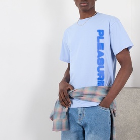 Model wearing Pleasures Buzzed Logo T-Shirt in Sky, with vertical blue logo print, styled with a plaid shirt tied at the waist and blue jeans.