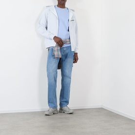Model wearing Pleasures Buzzed Logo Sky T-Shirt, styled with a white zip jacket, light wash jeans, and grey trainers.